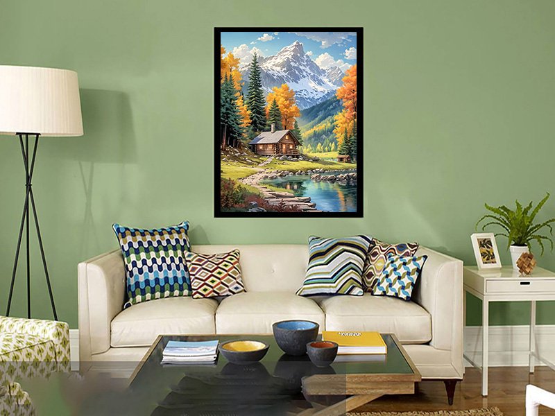 Diamond painting kit of a log cabin by a lake with mountains in the background, displayed on a wall above a sofa.
