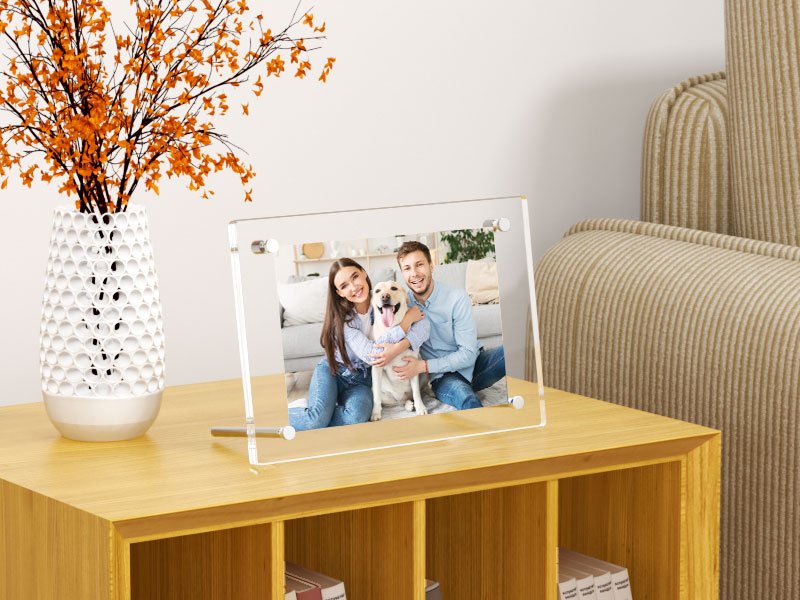 Clear acrylic picture frame displaying a smiling couple and a dog on a wooden table next to a vase of orange flowers.