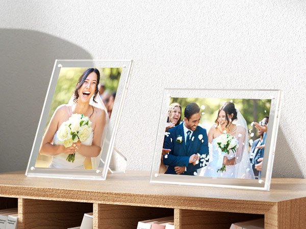 Two clear acrylic picture frames displaying wedding photos on a wooden shelf.