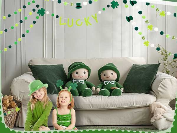 Two children and two plush dolls in a living room decorated for St. Patrick's Day with green and white garlands.