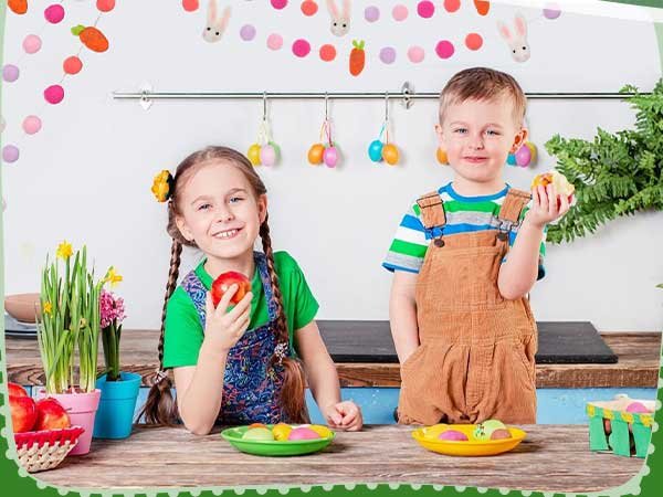Two smiling children holding apples at a table decorated with Easter-themed garlands and hanging eggs.