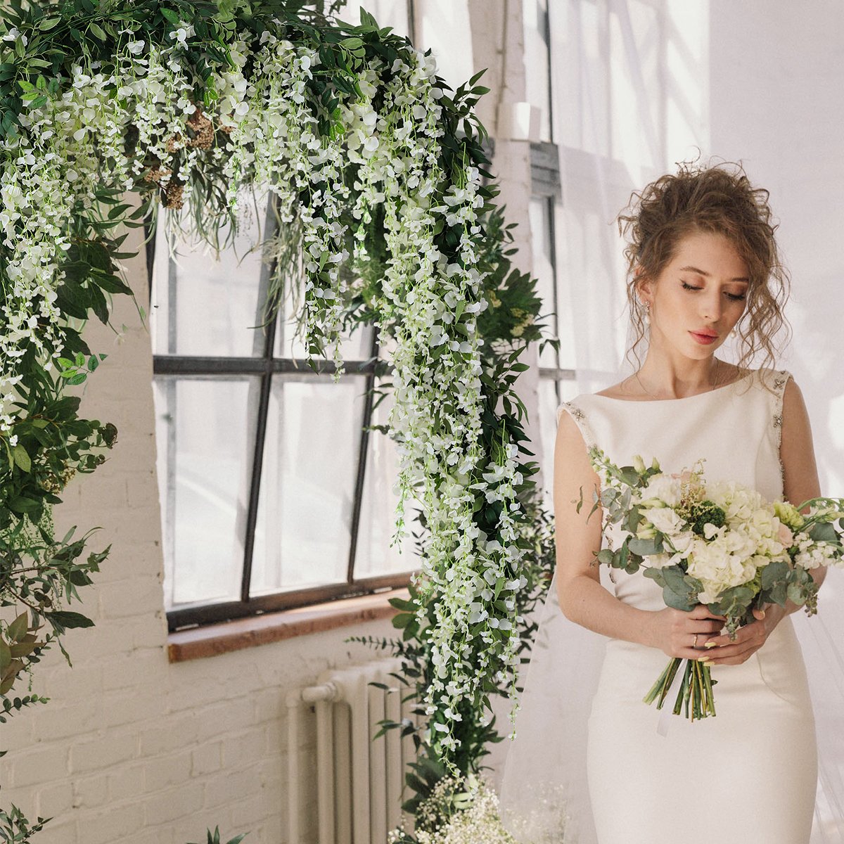 Bride holding a bouquet next to a wedding arch decorated with white wisteria vines and greenery