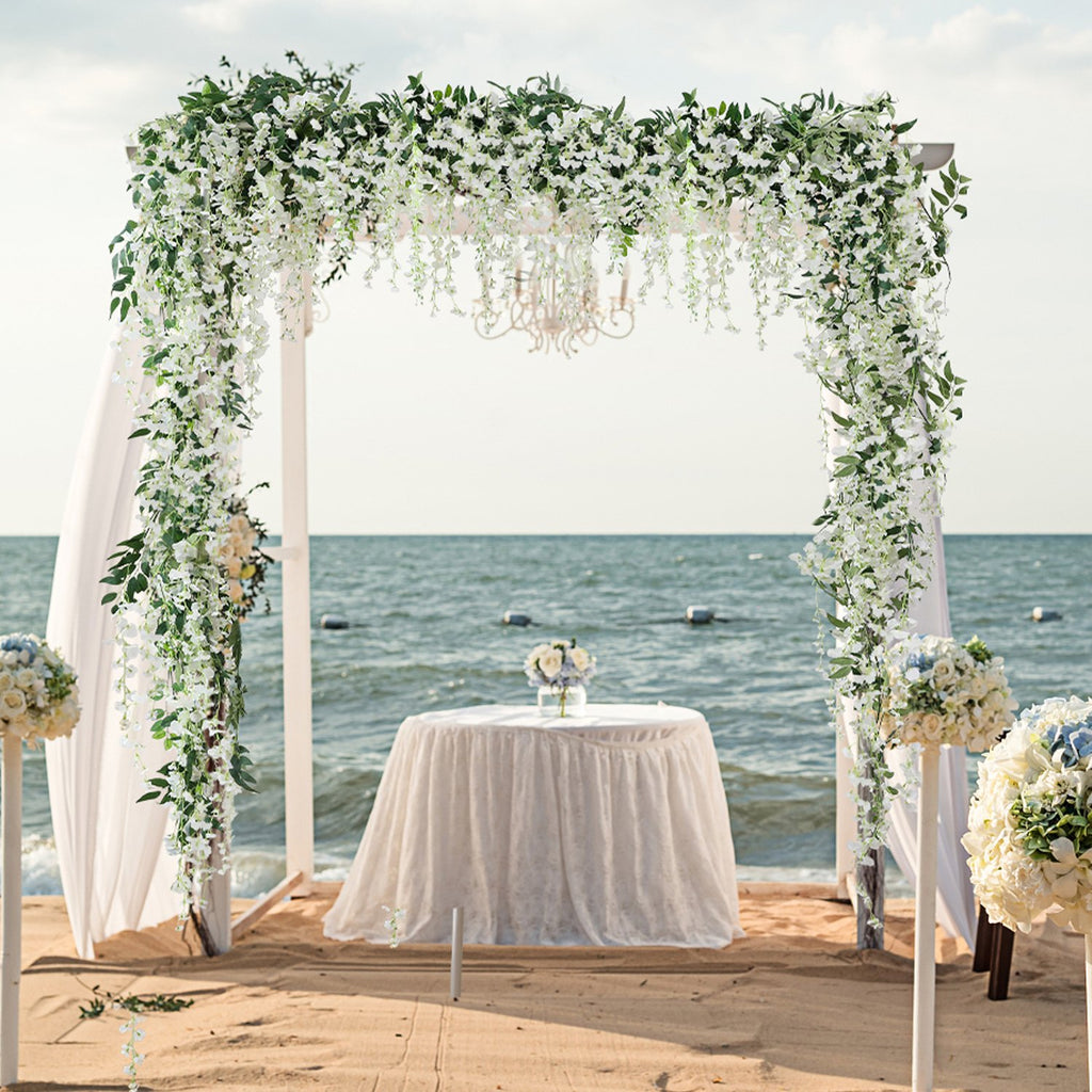 Wedding arch decorated with white wisteria vines and greenery on a beach with the ocean in the background.