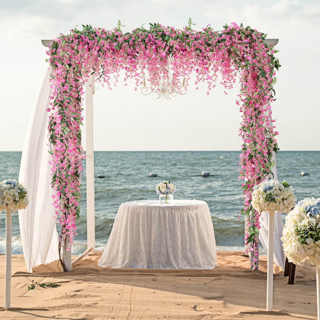 Wedding arch decorated with pink wisteria vines and greenery on a beach with a table and chairs set up.