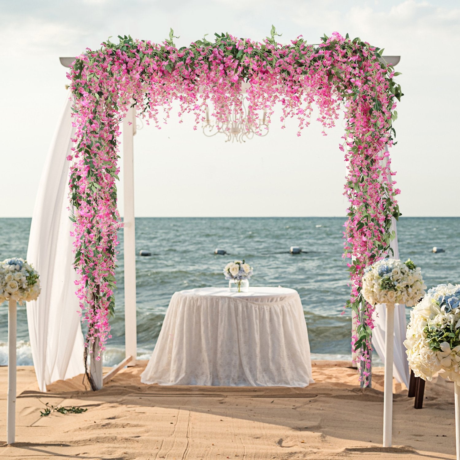Wedding arch decorated with pink wisteria vines and greenery on a beach with a table and chairs set up.