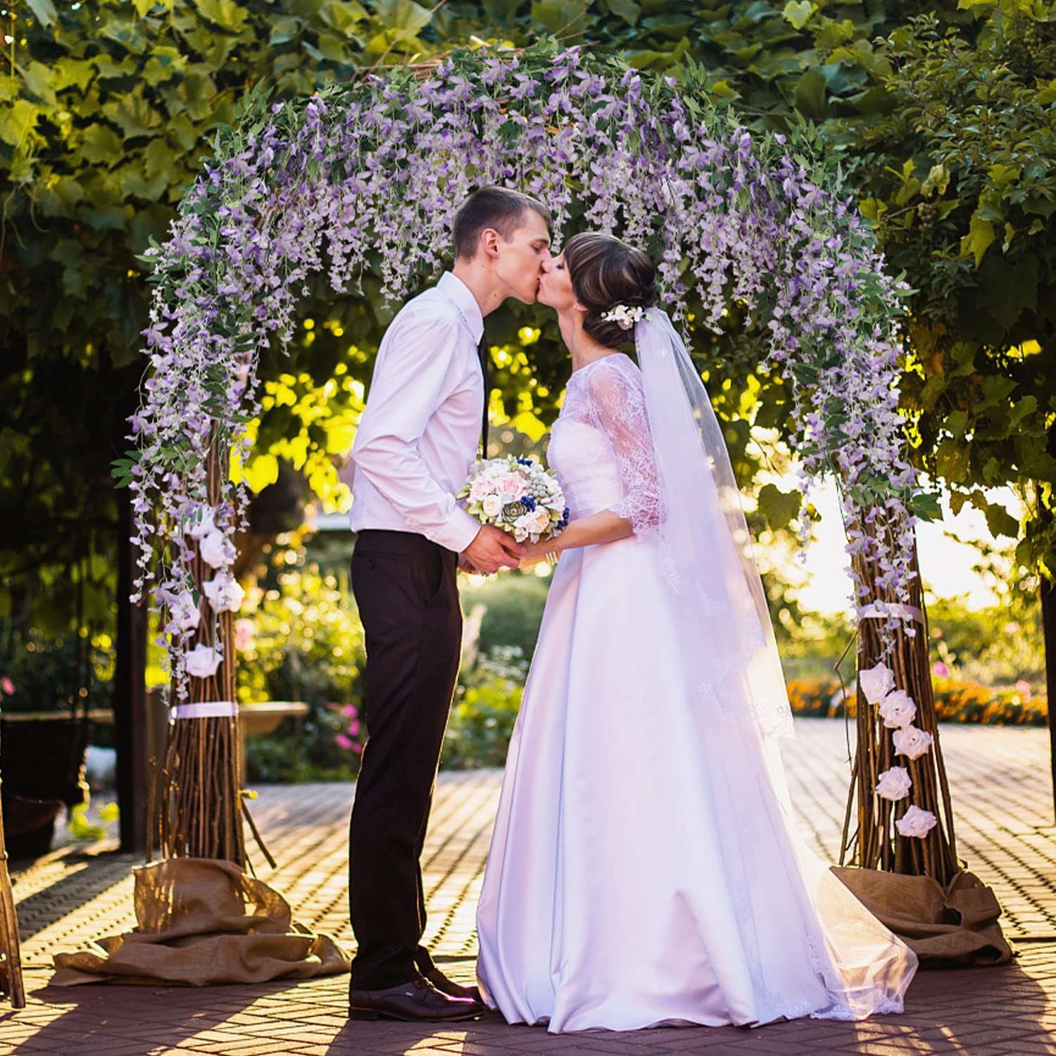 Bride and groom kissing under a wedding arch decorated with artificial purple wisteria vines and white roses.