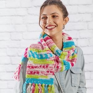 Woman smiling, wearing a colorful crocheted scarf with tassels against a white brick wall.