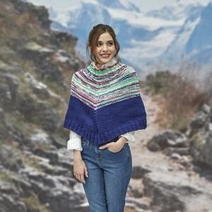 Woman wearing a colorful knitted poncho with blue trim, standing outdoors with a mountain backdrop.