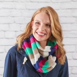 Woman wearing a colorful striped infinity scarf knitted from Aran yarn, against a white brick wall.