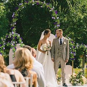 Wedding ceremony with a bride and groom standing under a floral arch decorated with purple roses and greenery.