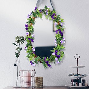 Artificial purple rose and green leaf garland draped around an oval mirror on a white wall.