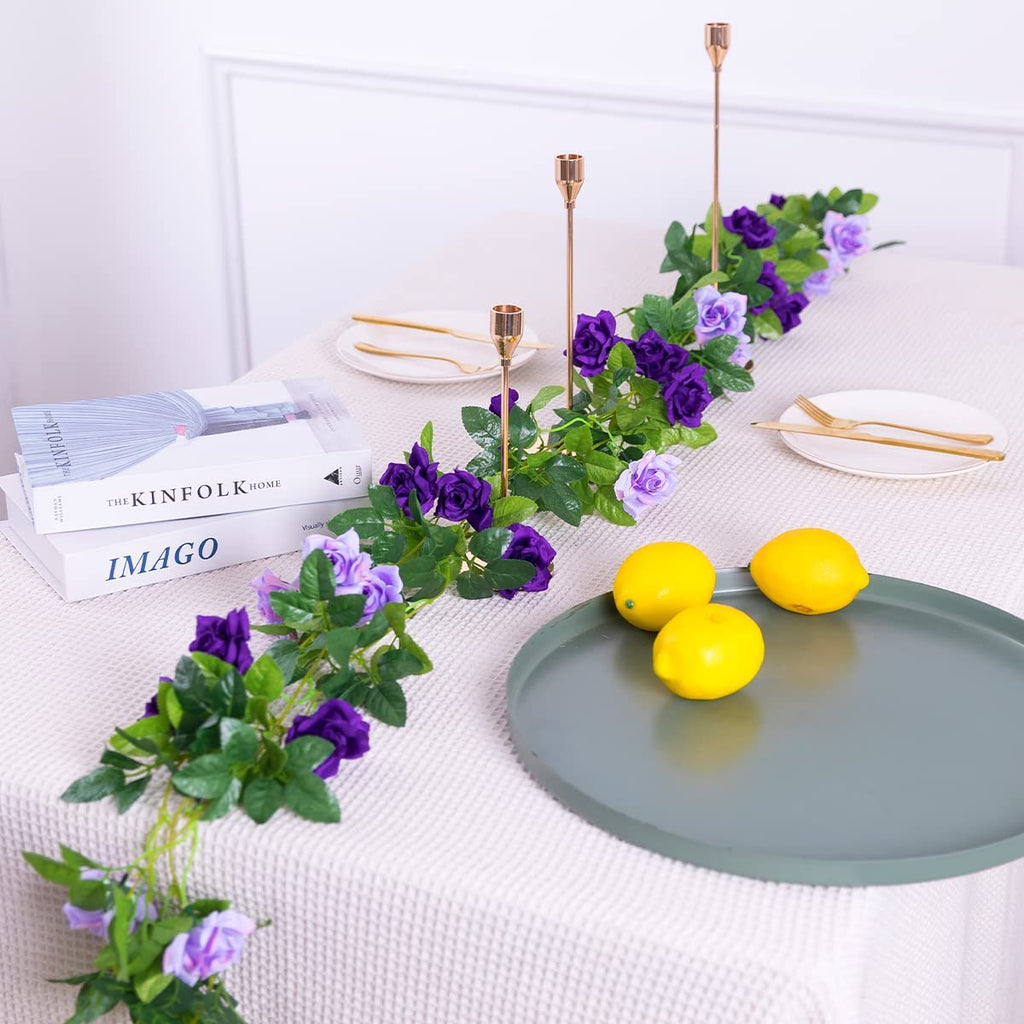 Artificial purple and lavender rose garland draped across a table with books, lemons, and candle holders.