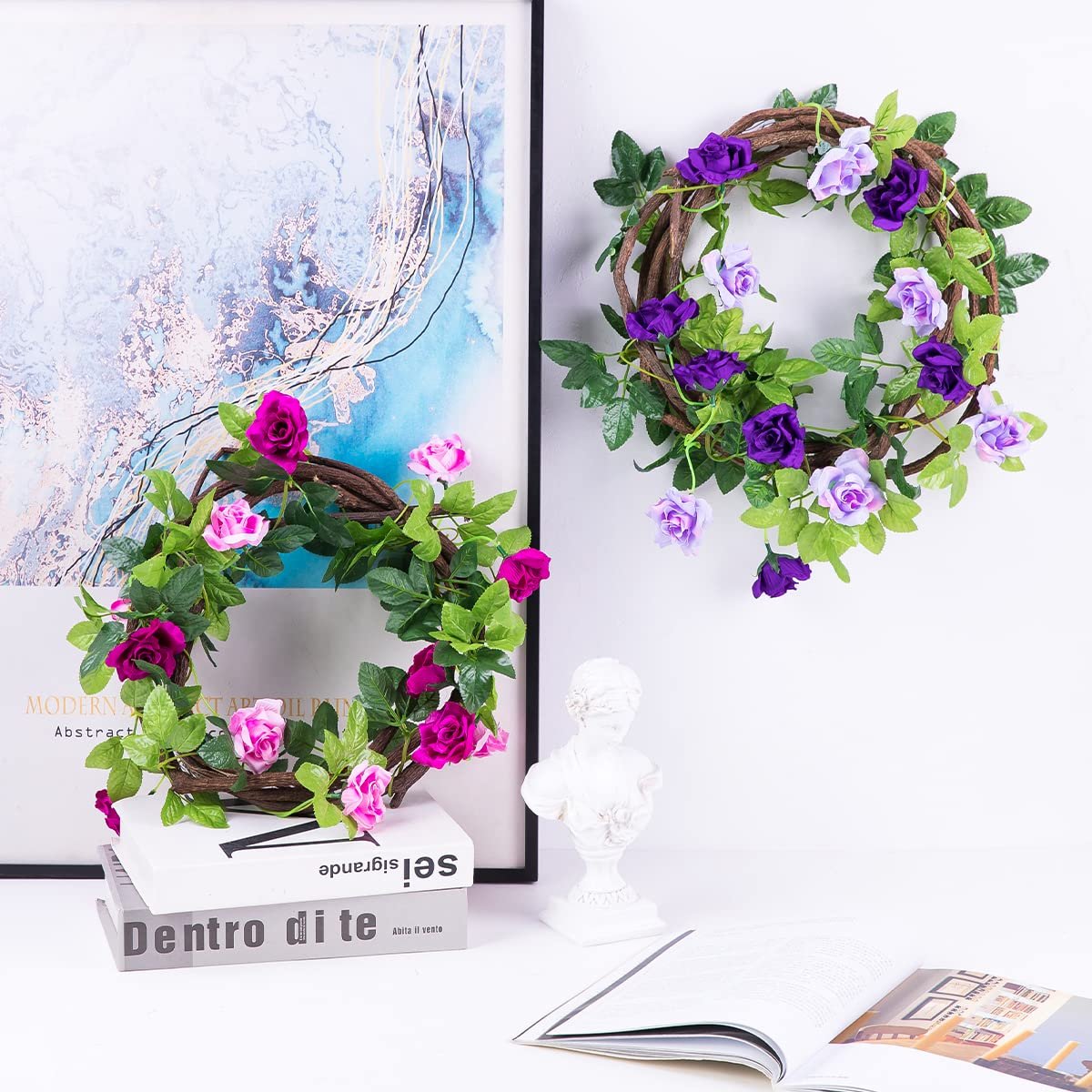 Two artificial rose garlands with purple and pink flowers and green leaves, displayed on a shelf with books and a bust.