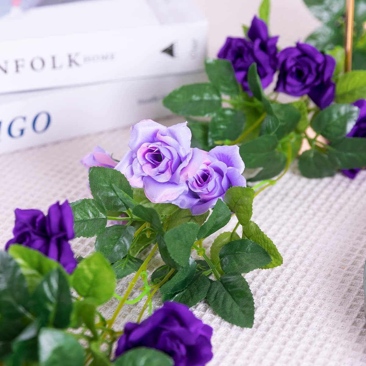 Artificial purple and lavender rose flower garland with green leaves on a textured white surface, with books in the background.