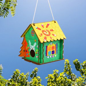 Decorated wooden birdhouse craft kit hanging outdoors against a blue sky with green foliage.