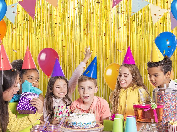Children celebrating a birthday party with a cake, balloons, and gold fringe curtains in the background.