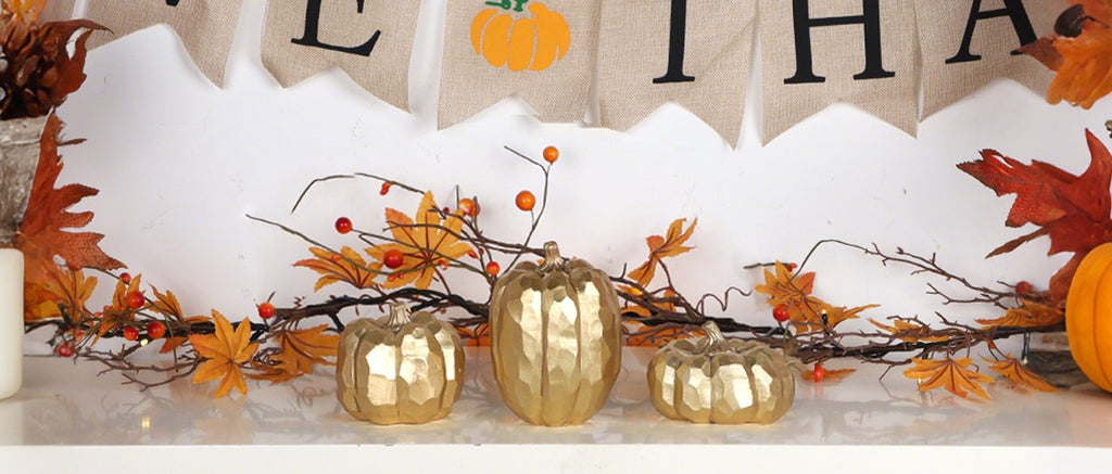 Three gold resin pumpkins with autumn leaves and berries on a white surface, with a burlap banner above.