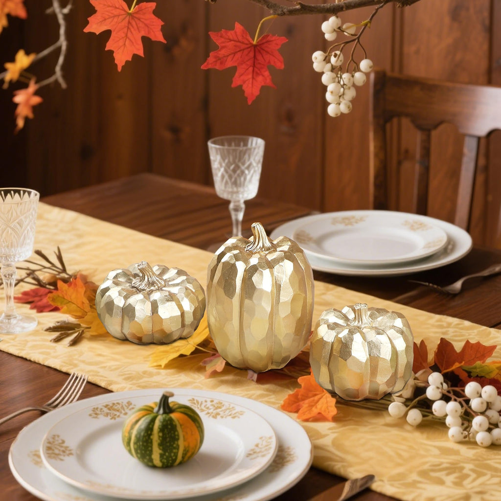 Three gold resin pumpkins and one small striped gourd arranged on a table runner with autumn leaves and berries.