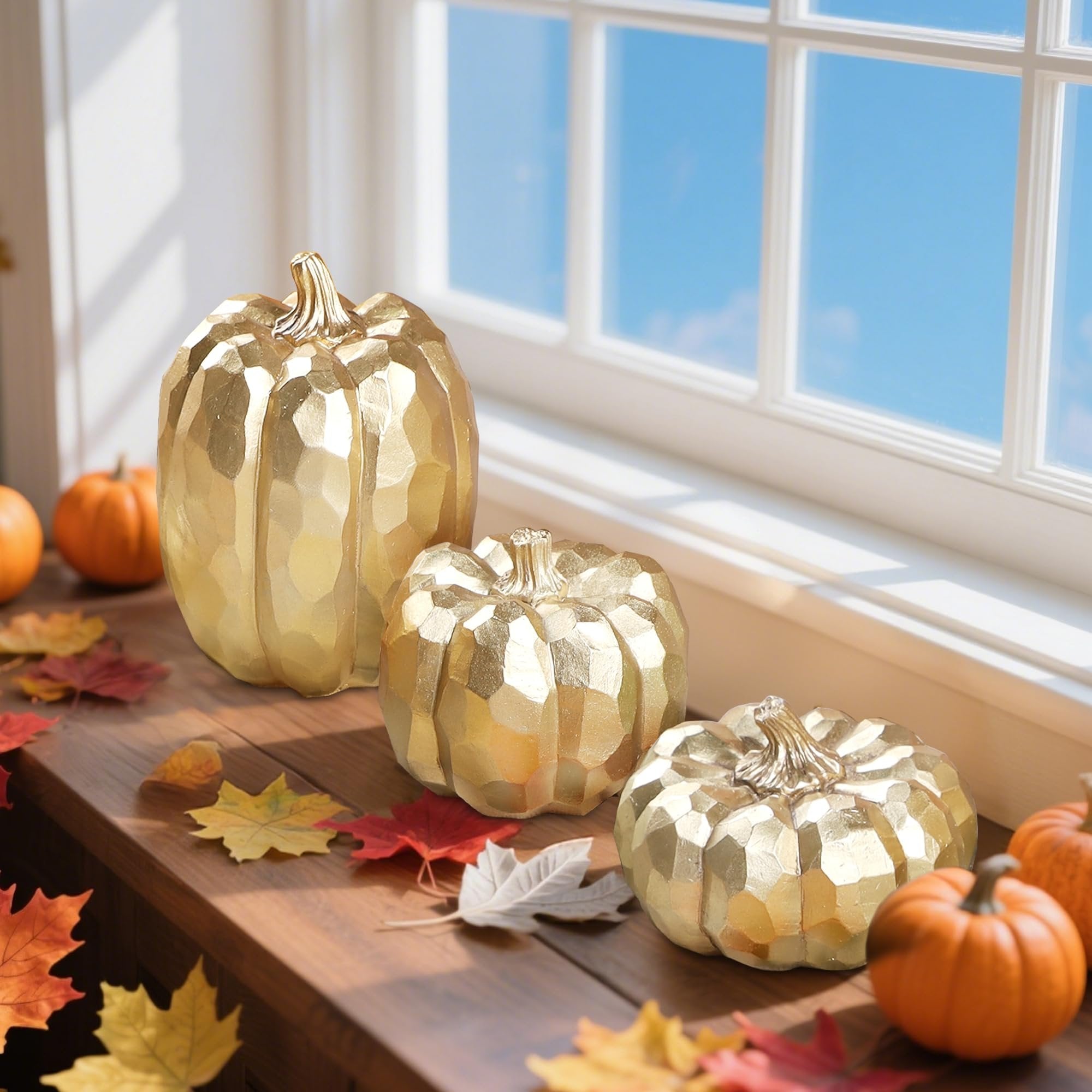Three gold faceted pumpkins and two small orange pumpkins arranged on a wooden surface with autumn leaves, near a window with a blue sky.