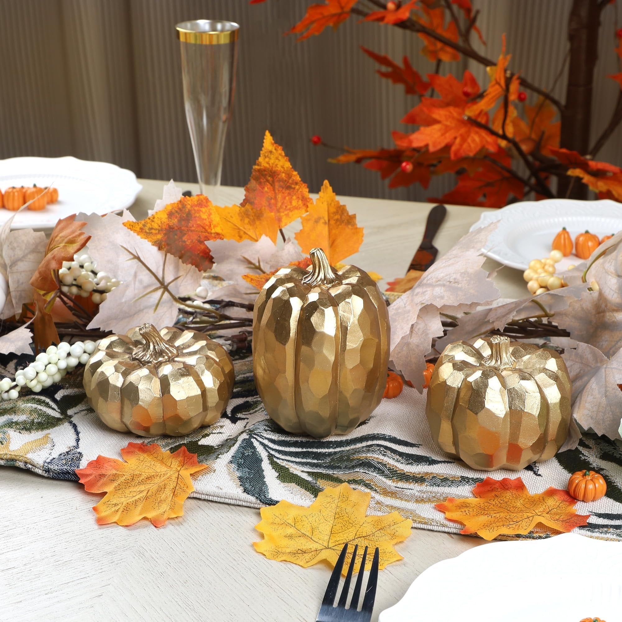 Three gold resin pumpkins arranged on a table runner with autumn leaves and place settings.