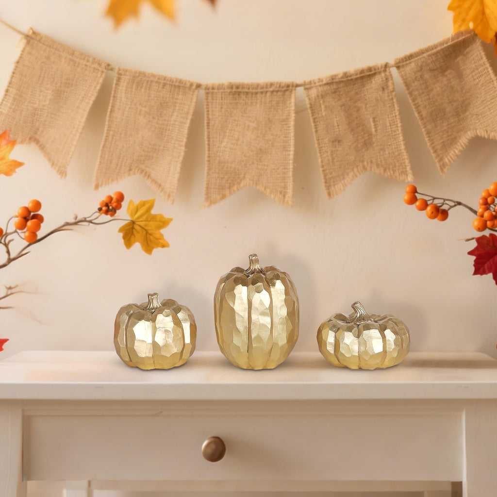 Three gold resin pumpkins of varying sizes displayed on a shelf with a burlap banner and autumn foliage in the background.
