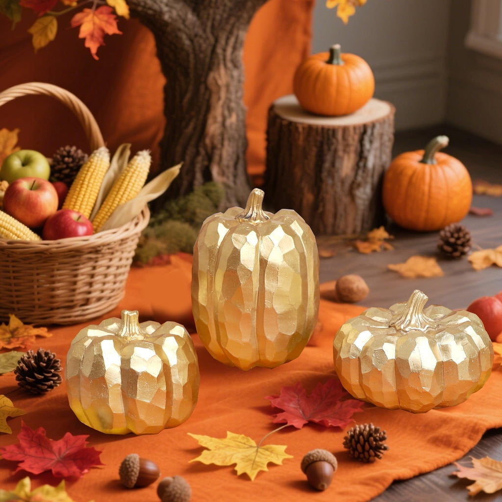 Three faceted gold pumpkins arranged on an orange cloth with autumn leaves, acorns, pinecones, and a basket of apples and corn in the background.
