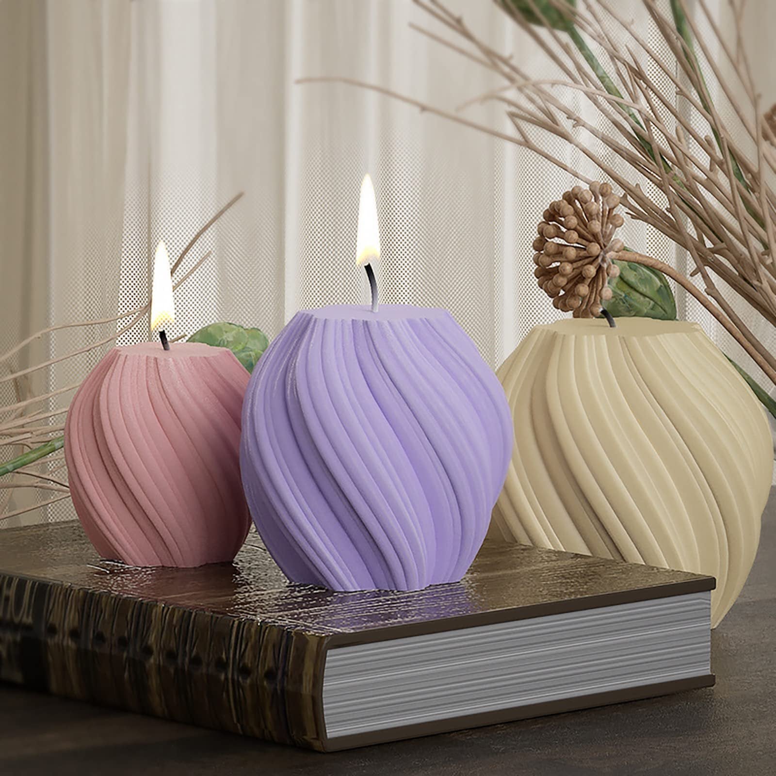 Three swirling candles in pink, purple, and cream colors sit on a book, with decorative branches in the background.