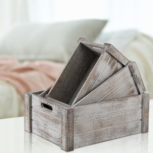Three rustic wooden crates with handles stacked on a white surface, with a blurred background of a bed and pillows.