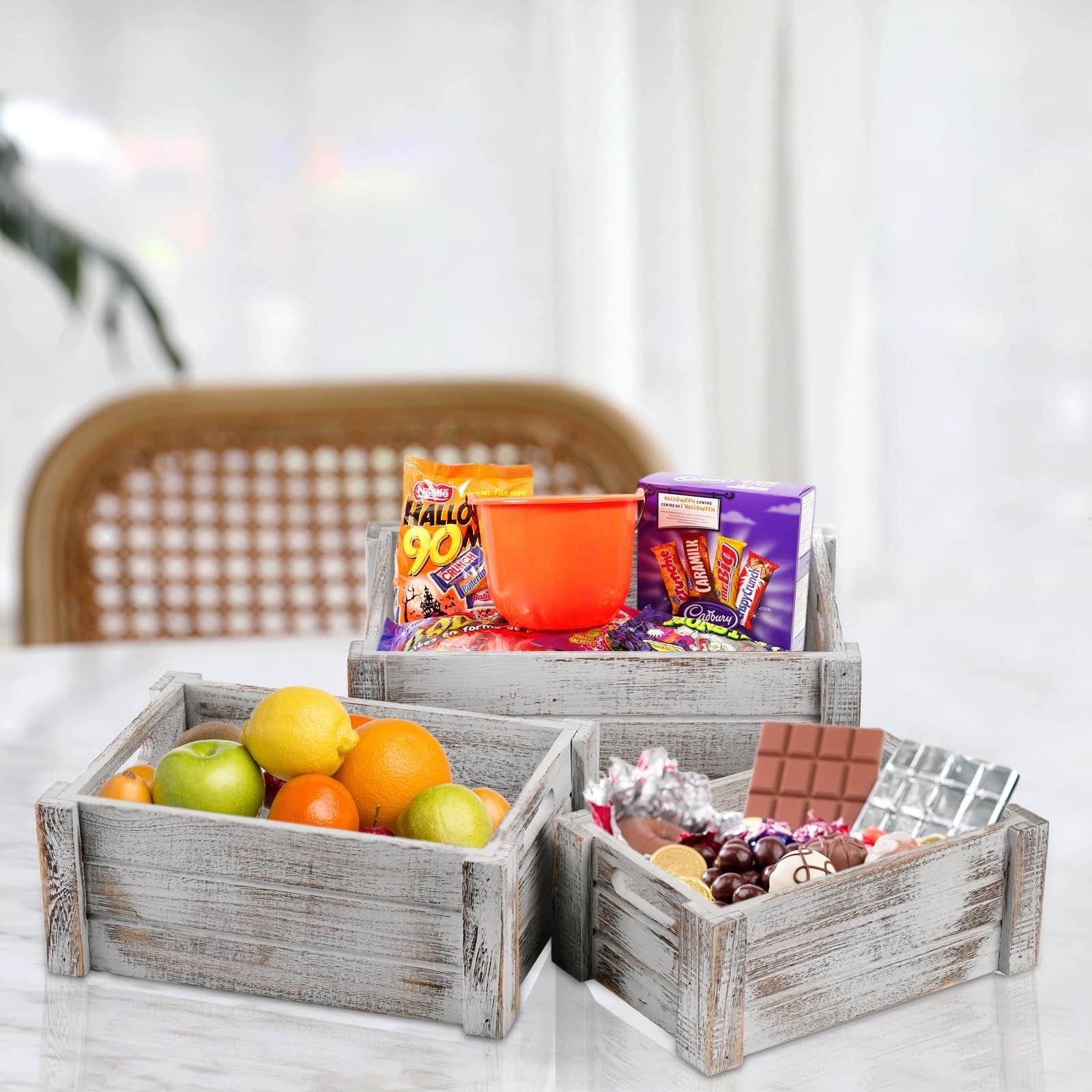 Three rustic wooden crates filled with fruit and candy on a marble surface. The crate in front contains apples, oranges, and lemons. The middle crate holds candy and a Halloween bucket. The back crate is filled with more candy.