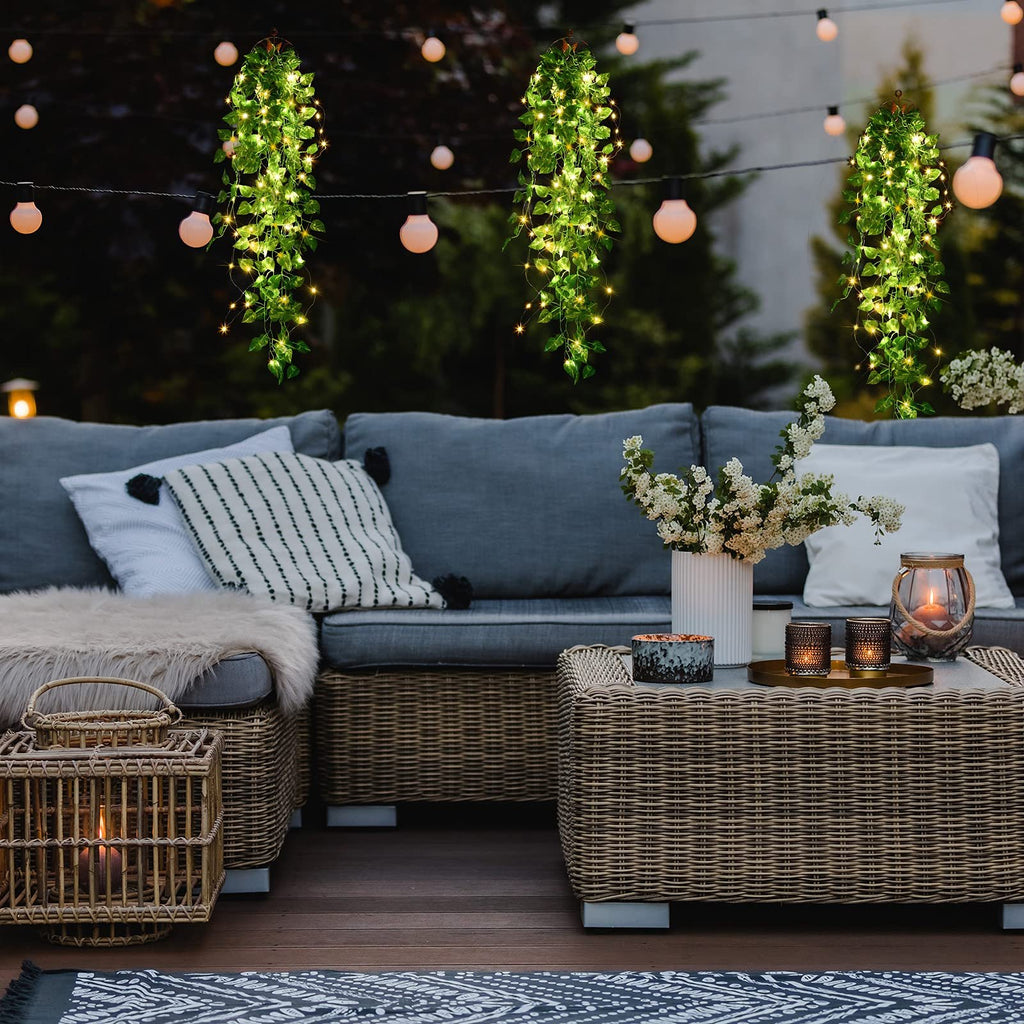 Three hanging artificial ivy vines with fairy lights draped over an outdoor patio sofa and coffee table at dusk.