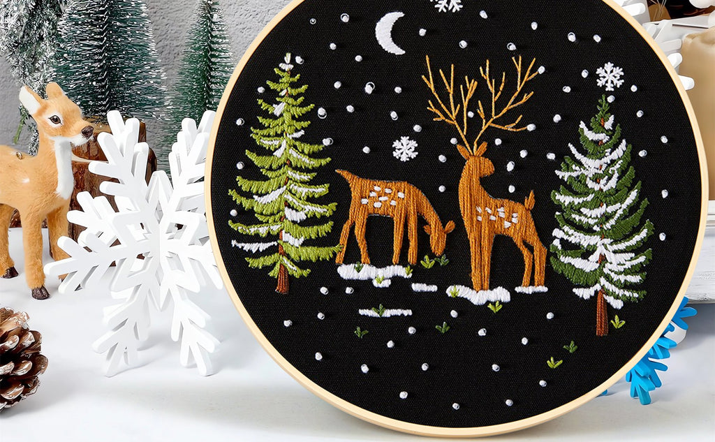 Embroidery hoop with a winter scene featuring deer and pine trees, surrounded by decorative elements.