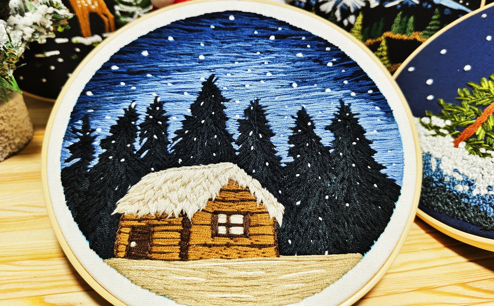 Embroidery hoop with a winter cabin scene, featuring dark pine trees under a starry blue sky with falling snow.