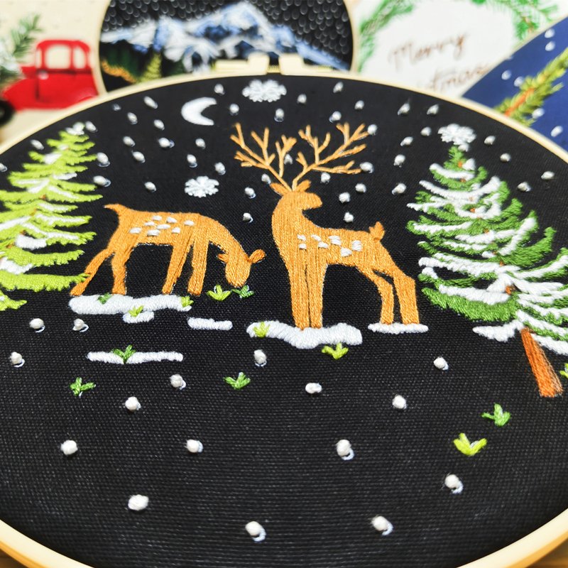 Embroidery hoop with a black fabric showing a winter scene with two deer, snow-covered pine trees, and falling snow.