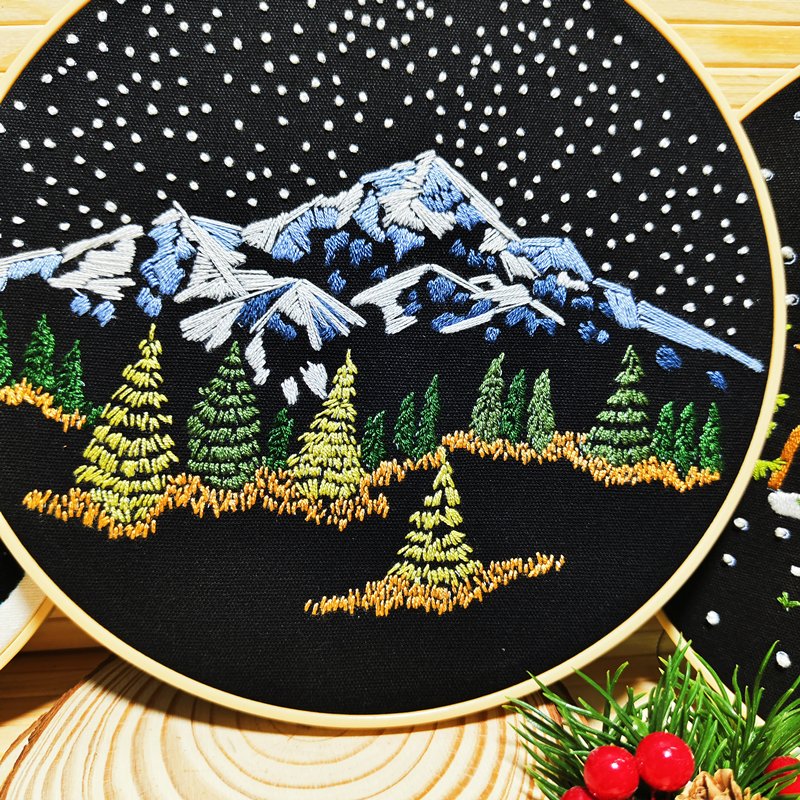 Embroidery hoop with a winter mountain landscape design featuring snow-capped peaks, pine trees, and falling snow.