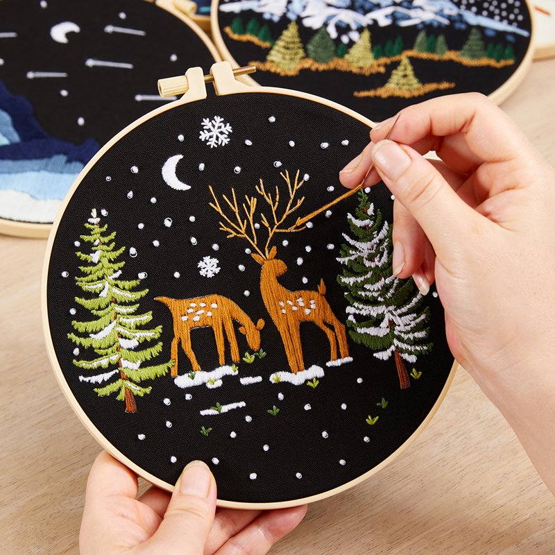 Embroidery hoop with a winter scene of two deer, pine trees, and snowflakes, being stitched by hands.