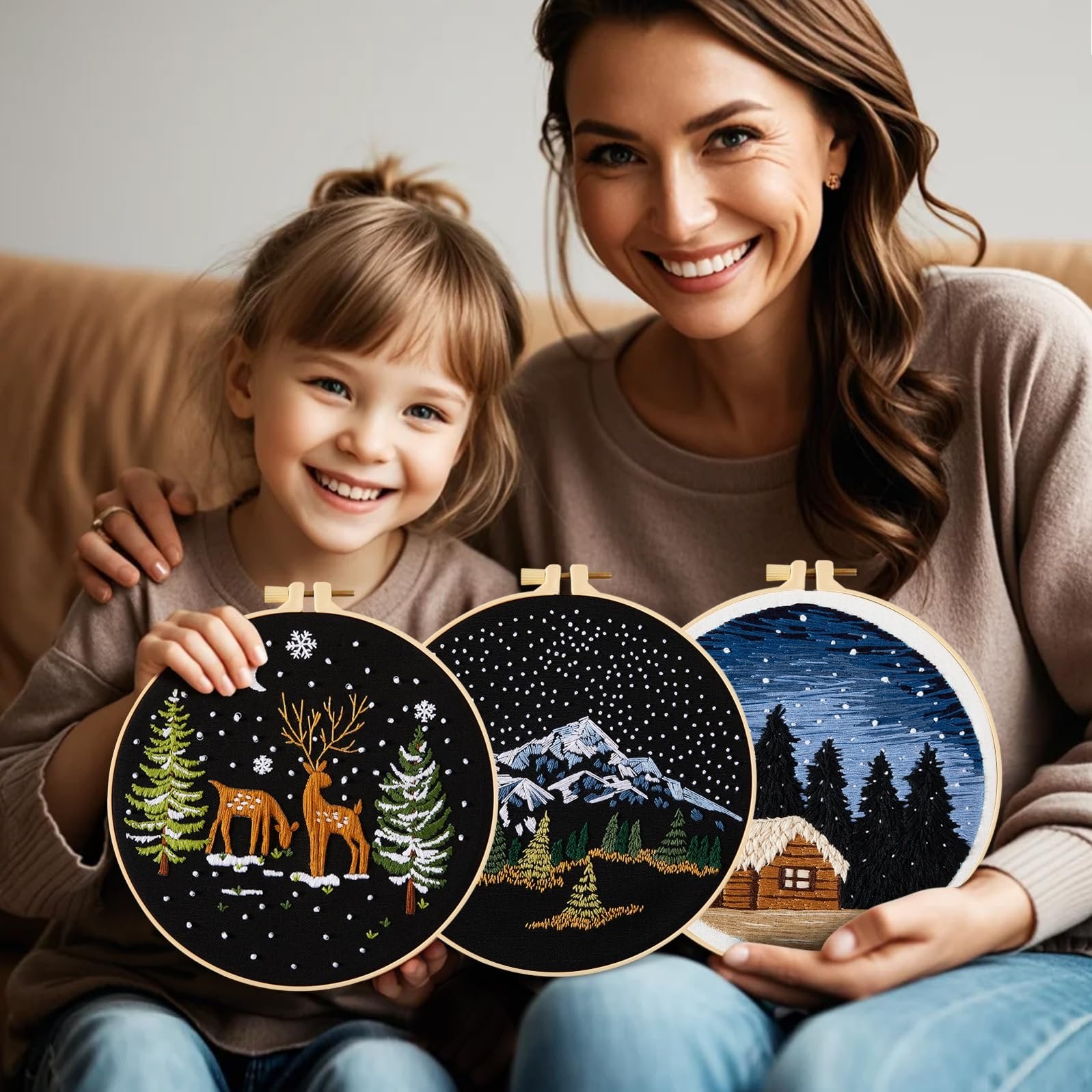Woman and child smiling, holding three embroidered hoops with winter scenes: deer and trees, mountains, and a cabin.