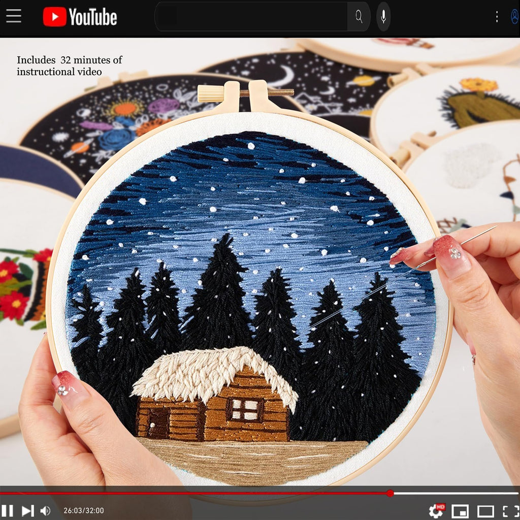 Embroidery hoop with a winter cabin and pine trees design being stitched by a hand. Other hoops with designs are visible in the background.