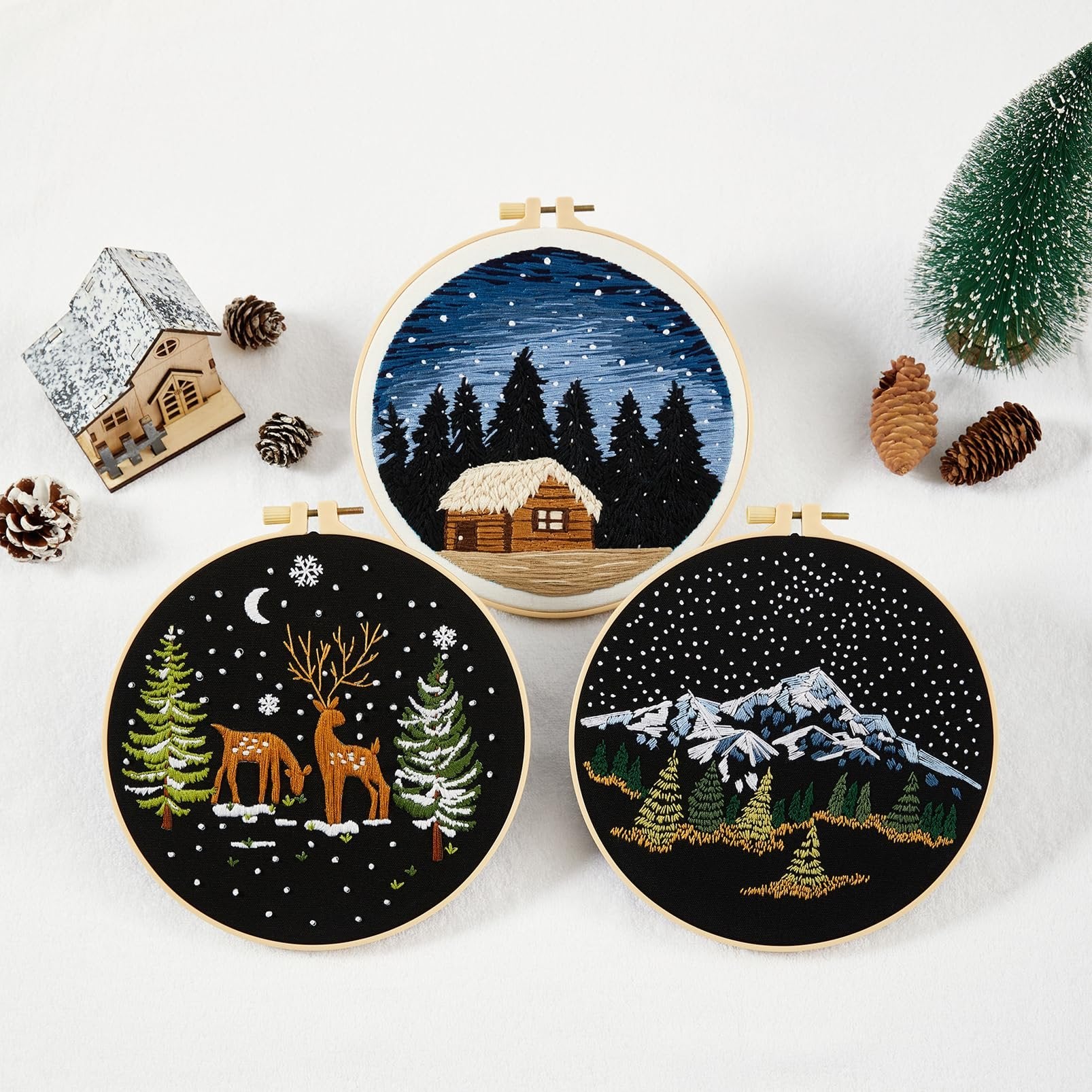 Three circular embroidery hoops with winter scenes: a cabin in snowy woods, deer and trees, and snowy mountains.
