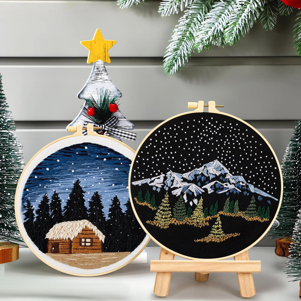 Two embroidered hoops showing winter scenes: one with a log cabin in a snowy forest, the other with snow-capped mountains and pine trees under a starry sky.