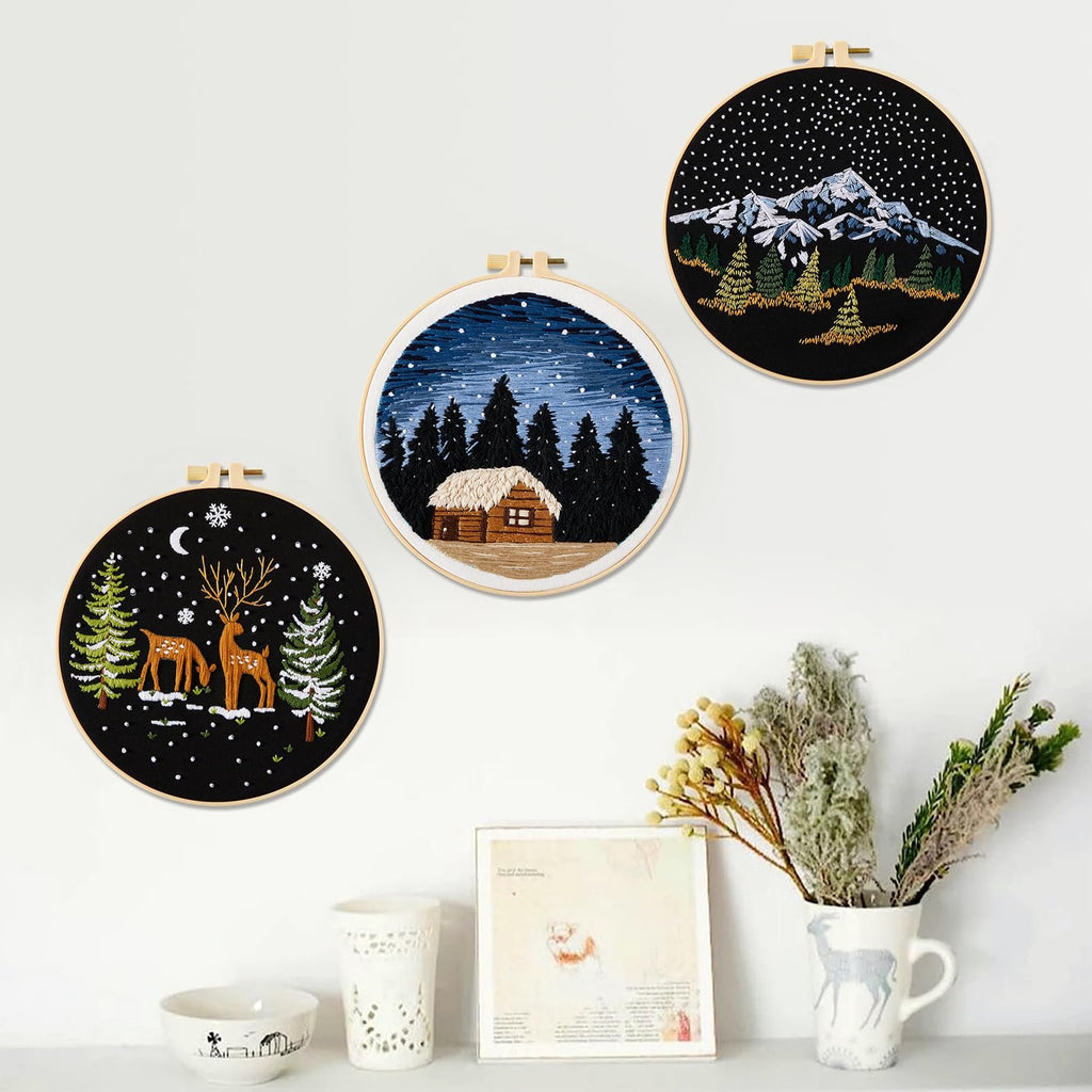 Three embroidered hoops depicting winter scenes: a cabin in a snowy forest, deer and trees under a crescent moon, and a mountain landscape under a starry sky.