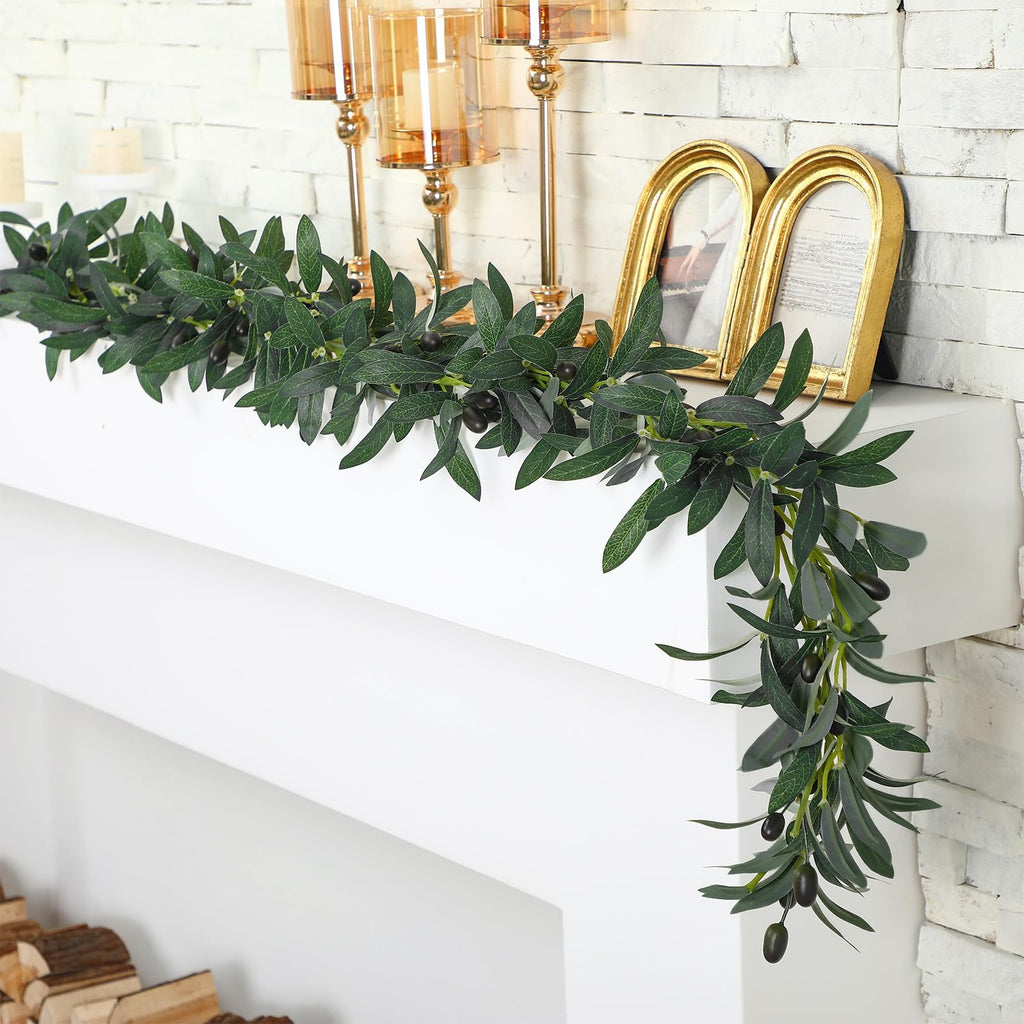 Artificial olive leaf garland with black olives draped over a white mantelpiece with gold picture frames and candle holders.