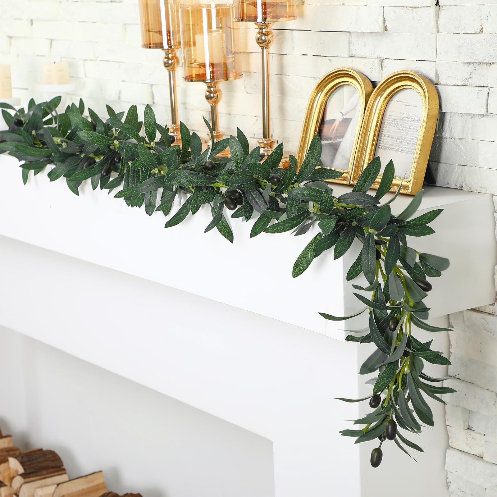 Artificial olive leaf garland with black olives draped over a white mantelpiece with gold picture frames and candle holders.
