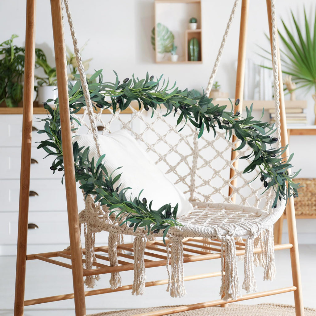 Artificial olive leaf garland with black olives draped over a macrame swing chair with a white pillow
