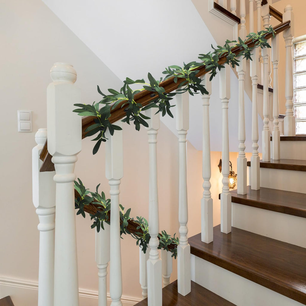 Artificial olive leaf garland with faux fruits draped along a wooden banister on a staircase.