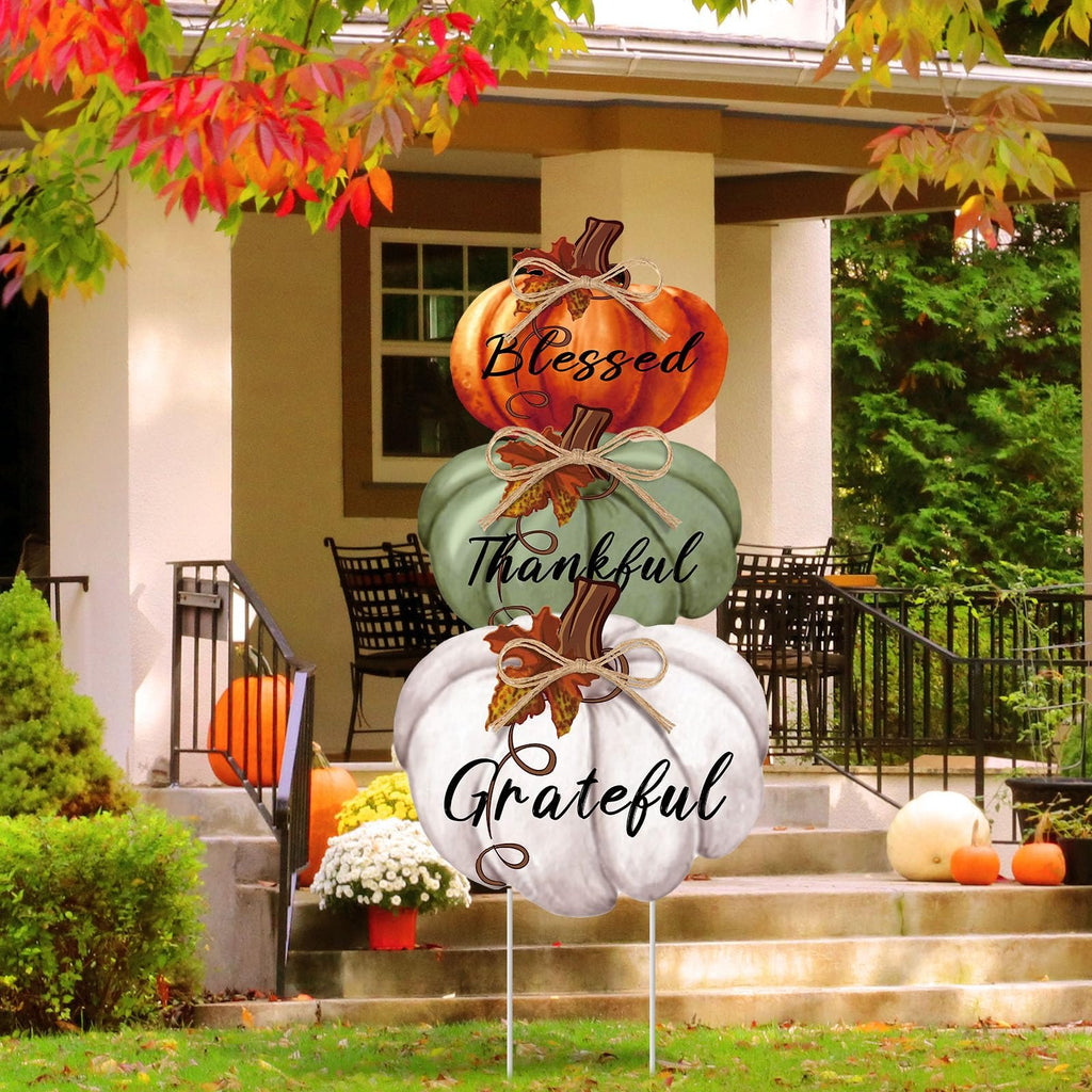 Stackable Thanksgiving pumpkin yard sign with Blessed, Thankful, Grateful text, displayed on a porch with autumn foliage.