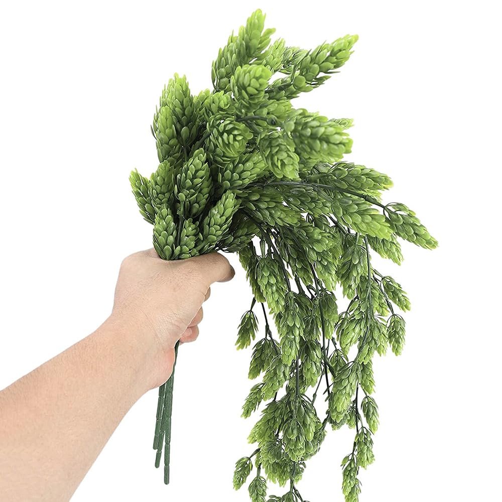 Hand holding a bunch of artificial green hops vines with multiple branches and cone-shaped blooms against a white background.
