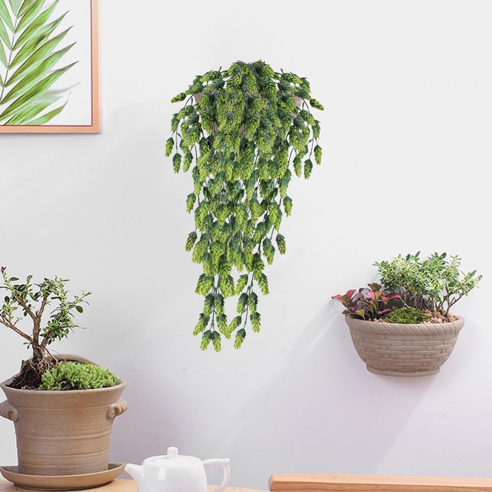 Artificial hops vine with green leaves and cones hanging on a white wall, with potted plants and a teapot below.