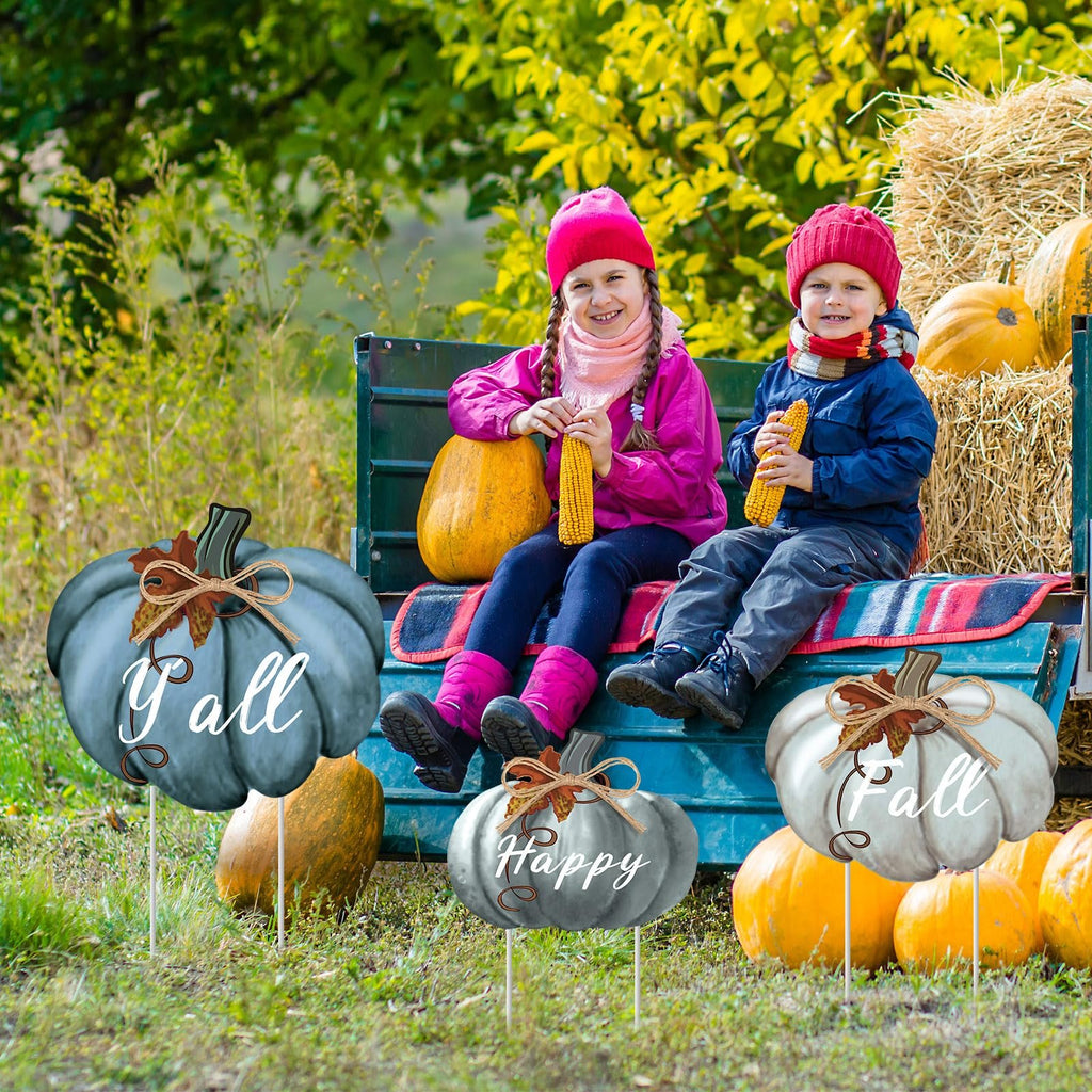 Two children sit on a truck bed with pumpkins and hay bales, surrounded by three pumpkin-shaped yard stakes with fall greetings.