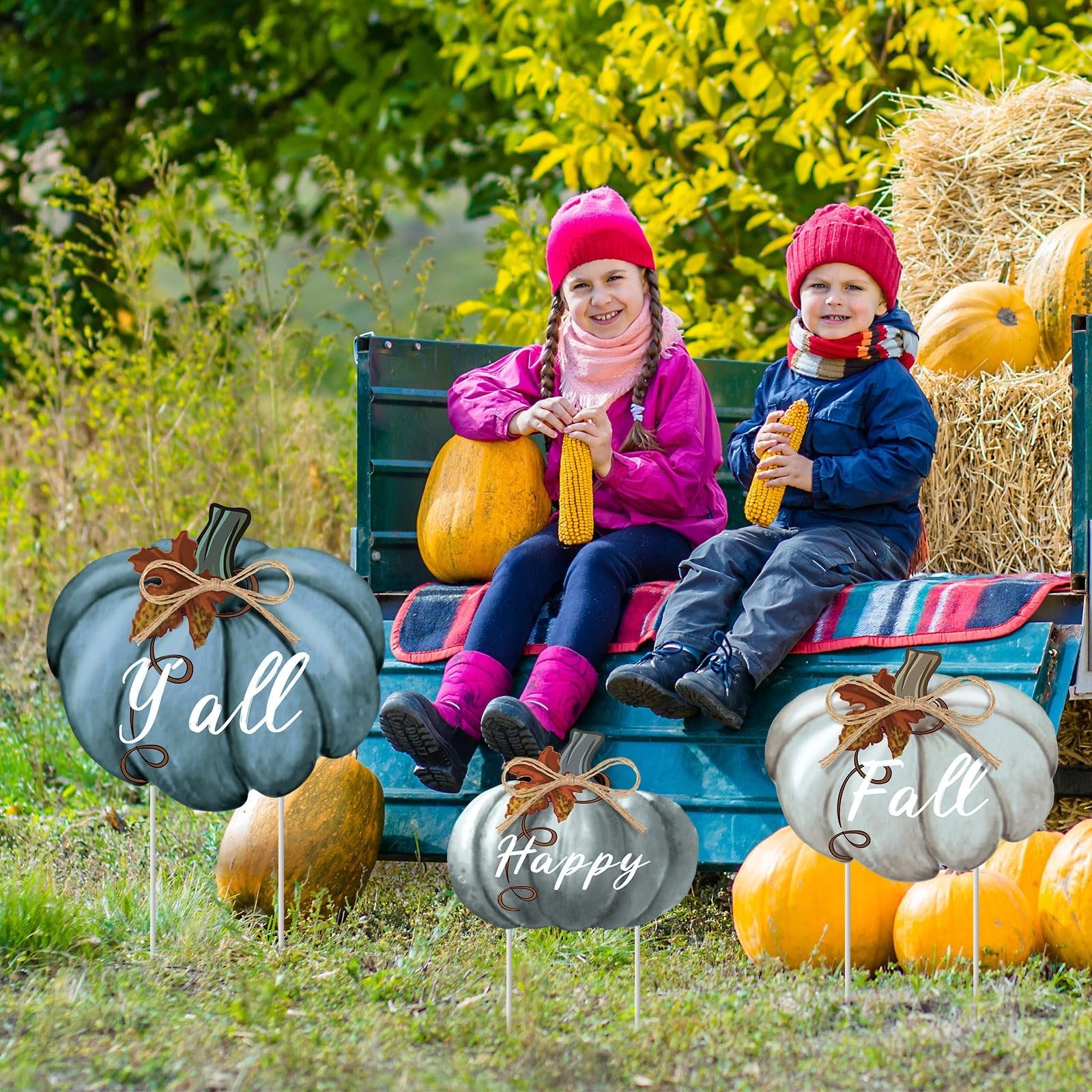 Two children sit on a truck bed with pumpkins and hay bales, surrounded by three pumpkin-shaped yard stakes with fall greetings.