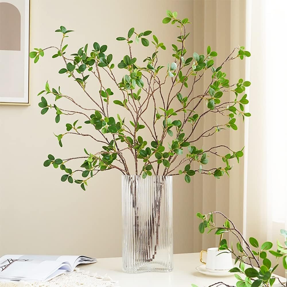 Artificial eucalyptus branches with green leaves in a clear ribbed vase on a table with an open book and a teacup.